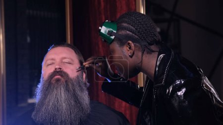 Téléchargez les photos : Une styliste afro-américaine en cuir noir peigne la barbe d'un client dans un salon de coiffure - en image libre de droit
