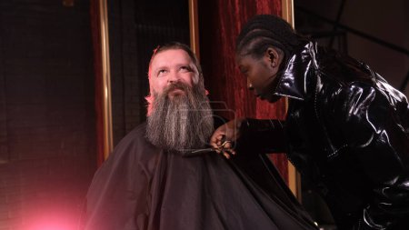Téléchargez les photos : Une styliste afro-américaine nivelle la barbe d'un homme avec des ciseaux dans un salon de coiffure. - en image libre de droit