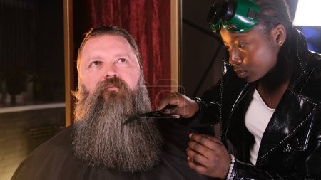 Téléchargez les photos : Une coiffeuse afro-américaine peigne et coupe la barbe d'un homme - en image libre de droit