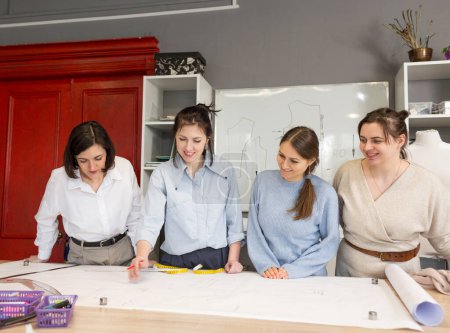 Photo for The concept of small business. Four female fashion designers work together in a tailoring studio. A group of women dressmakers discusses fabric, pattern and sketch in a sewing workshop. - Royalty Free Image