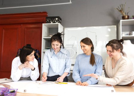 Photo for Four female fashion designers work together in a tailoring studio. A group of women dressmakers discusses fabric, pattern and sketch in a sewing workshop. The concept of small business - Royalty Free Image