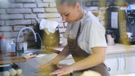 Téléchargez les photos : Une femme dans une cuisine ouverte dans un restaurant déploie la pâte avec un rouleau à pâtisserie pour faire des saucisses dans la pâte - en image libre de droit