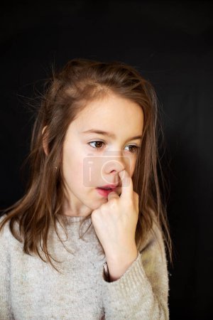 Foto de Una chica desaliñada con un dedo en la nariz sobre un fondo oscuro. - Imagen libre de derechos