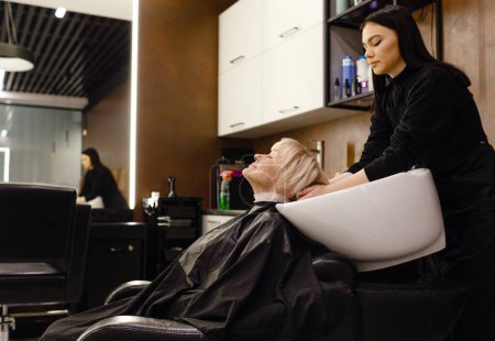 Téléchargez les photos : Portrait d'une femme âgée visitant un coiffeur professionnel. Un coiffeur expérimenté lave les cheveux du client - en image libre de droit