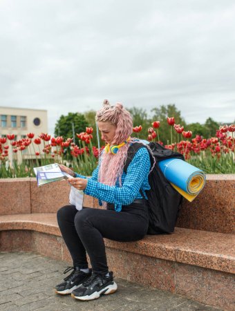 Foto de Un atractivo turista joven con una camisa azul está explorando una nueva ciudad. Una chica con el pelo largo y rosado sostiene un mapa de papel del esquema de la ciudad en sus manos. Viajar por Europa - Imagen libre de derechos