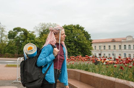 Téléchargez les photos : Une fille avec des écouteurs et des cheveux roses voyage autour de la ville avec un sac à dos sur les épaules. Parler au téléphone. - en image libre de droit