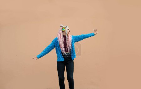 Photo for An attractive young woman with pink long dreadlocked hair and a blue shirt, rhythmically dancing to music with headphones on a beige background. Empty space for advertising text. - Royalty Free Image