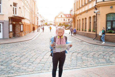Foto de Un atractivo turista joven con una camisa azul está explorando una nueva ciudad. Una chica con el pelo largo y rosado sostiene un mapa de papel del esquema de la ciudad en sus manos. Viajar por Europa - Imagen libre de derechos