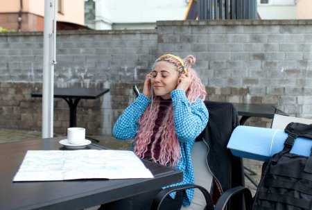 Foto de Un atractivo turista joven en una camisa azul bebe café y escucha música con auriculares. Una chica con el pelo largo y rosado está almorzando en un café. Viajar por Europa - Imagen libre de derechos