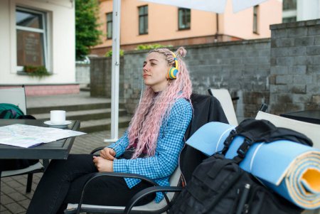 Foto de Un atractivo turista joven en una camisa azul bebe café y escucha música con auriculares. Una chica con el pelo largo y rosado está almorzando en un café. Viajar por Europa - Imagen libre de derechos