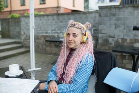Photo for An attractive young tourist in a blue shirt drinks coffee and listens to music with headphones. A girl with long pink hair is having lunch in a cafe. Travel around Europe - Royalty Free Image