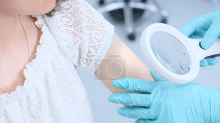 Foto de Una dermatóloga usando una lupa médica en la clínica examina la piel del paciente - Imagen libre de derechos