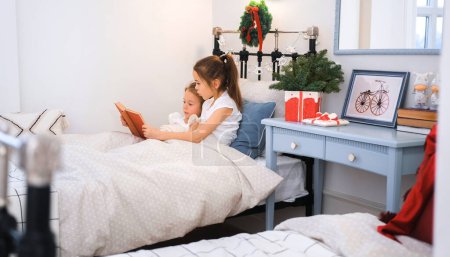 Photo for Two little sisters are reading a book in bed while in a room decorated with Christmas decorations and garlands - Royalty Free Image