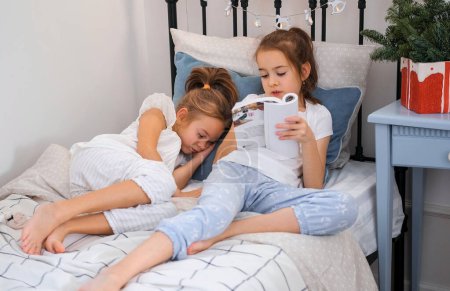 Foto de Una hermana mayor está leyendo un libro en la cama a su hermana menor que se ha quedado dormida. Noche de Navidad. - Imagen libre de derechos