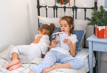 Téléchargez les photos : Une sœur aînée lit un livre au lit à sa sœur cadette qui s'est endormie. Nuit de Noël. - en image libre de droit