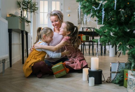 Téléchargez les photos : Maman heureuse étreint ses petites filles assises sur le sol près du sapin de Noël - en image libre de droit