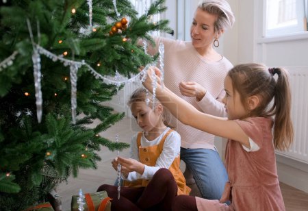 Téléchargez les photos : Maman et ses filles décorent un sapin de Noël assis sur le sol dans le salon - en image libre de droit