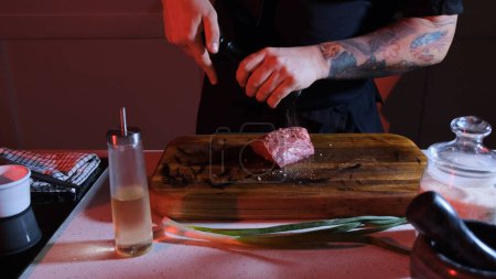 Foto de El chef espolvorea un trozo de carne cruda con pimienta. Ambiente de trabajo en la mesa de la cocina. - Imagen libre de derechos