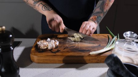 Téléchargez les photos : Le cuisinier hache finement l'ail sur une planche à découper en bois dans la cuisine - en image libre de droit