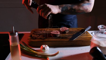 Téléchargez les photos : Un chef masculin saupoudrer de poivre sur le steak de bœuf porterhouse dans la cuisine du restaurant. un homme cuisine un steak - en image libre de droit