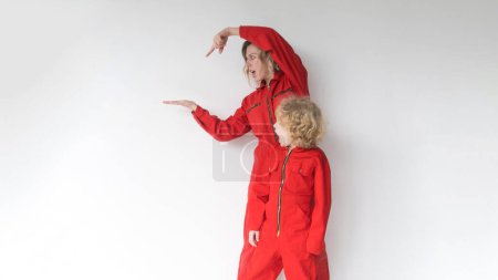 Eine positive Blondine mit einem kleinen Sohn in roten Jumpsuits zeigt auf die Stelle, an der Ihr Werbetext vor weißem Hintergrund angezeigt wird..