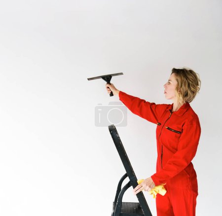 Téléchargez les photos : Une jeune fille en combinaison rouge, employée d'une entreprise de nettoyage, debout sur un escabeau et tenant un grattoir de verre à la main sur un fond blanc isolé - en image libre de droit