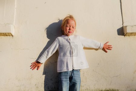 Téléchargez les photos : Une petite fille se tient contre un mur par une journée d'été ensoleillée dans un sweat à capuche blanc, fermant les yeux du soleil - en image libre de droit