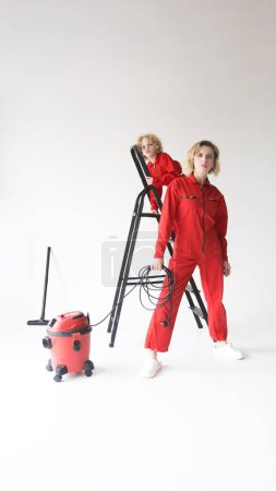 Téléchargez les photos : Une femme et un garçon en uniforme rouge d'entreprise de nettoyage sur fond blanc à côté d'un aspirateur. Nettoyage familial. - en image libre de droit