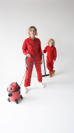 Téléchargez les photos : Maman et fils en salopette rouge sur un stand de fond isolé blanc à côté d'un thème de nettoyage et de nettoyage d'aspirateur - en image libre de droit