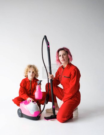 Téléchargez les photos : Un garçon et une femme adulte en salopette rouge sur fond blanc avec des appareils ménagers pour nettoyer la maison. - en image libre de droit