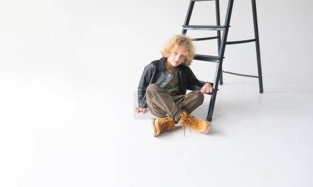 Foto de Un niño pequeño con el pelo rubio rizado se sienta casualmente en el suelo, apoyado en una escalera oscura. Llevan una chaqueta de cuero negro, pantalones marrones y botas amarillas, dando un toque caprichoso a su pose relajada.. - Imagen libre de derechos