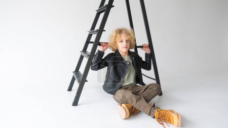A boy with blonde curly hair is sitting on the floor, looking at the camera, with a black ladder behind him in a neutral-toned studio. He is wearing a black leather jacket, an olive green shirt, brown trousers, and tan boots.