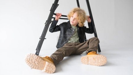 Photo for A young boy with voluminous curly hair sits nonchalantly underneath a black ladder, casting a gaze downward. He is wearing a black leather jacket, brown pants, and contrasting yellow boots, creating a relaxed yet stylish look. The ladder suggests an - Royalty Free Image