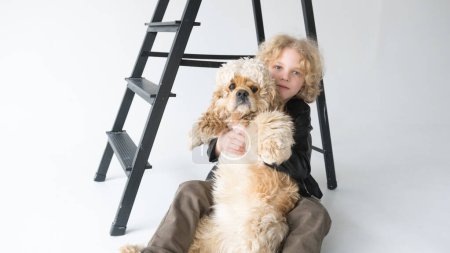 A young boy with curly blond hair is sitting on the floor leaning against a black step ladder while tightly hugging his large, fluffy, light-brown and white dog, both looking directly towards the camera.