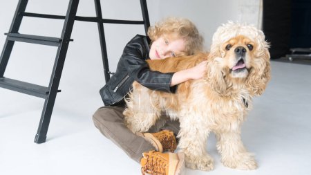 Photo for Young A boy with curly hair dressed in a black leather jacket and beige pants is gazing affectionately at his loyal companion, a large fluffy dog with a playful demeanor. They are sitting together against a white background, with a hint of a black la - Royalty Free Image