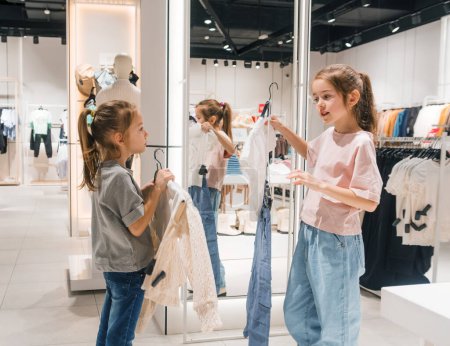 Téléchargez les photos : Deux petites filles regardent à travers les vêtements dans le magasin, explorant activement diverses options. - en image libre de droit