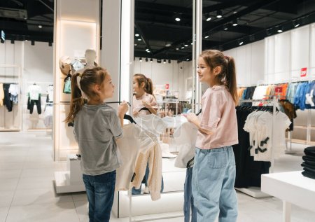 A pair of girls are choosing outfits and holding clothes up to themselves in a brightly lit clothing store.