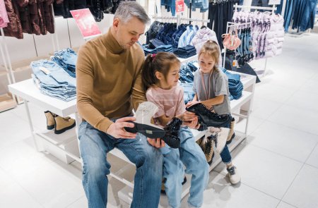 Photo for A dad and his girls sitting at a clothing store, trying on black shoes. - Royalty Free Image