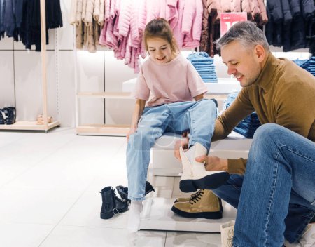 Téléchargez les photos : Une fille s'assoit pendant que son père aide à mettre de nouvelles chaussures sur ses pieds dans un magasin. - en image libre de droit