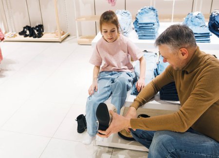 Une fille s'assoit pendant que son père aide à mettre de nouvelles chaussures sur ses pieds dans un magasin.
