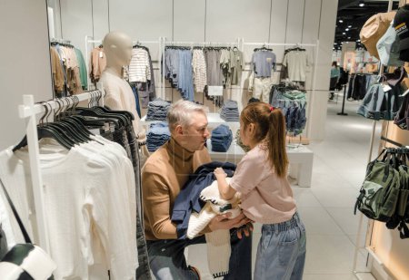 A father and daughter are selecting clothes together at a bustling retail store, surrounded by racks of clothing and accessories.