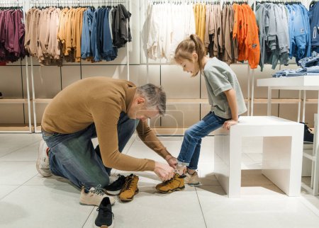 Un padre ayuda a su hija a probarse un par de zapatos deportivos marrones en una tienda de ropa bien iluminada y organizada..
