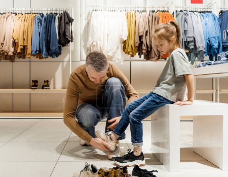 A father assists his daughter in trying on sneakers inside a clothing store filled with neatly arranged garments during the afternoon.
