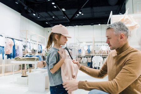 Téléchargez les photos : A father assists his young daughter with trying on clothes in a well-lit, contemporary clothing store. - en image libre de droit