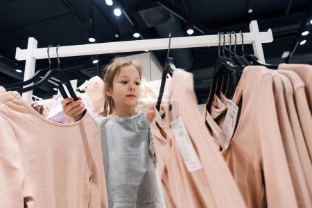 Ein junges Mädchen stöbert in einem modernen Bekleidungsgeschäft in einem Regal mit rosa Hemden und untersucht jedes Kleidungsstück sorgfältig..