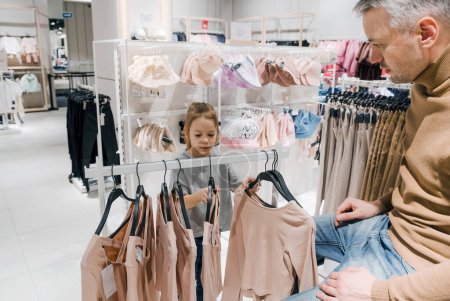 A man and a young girl browse and select clothes together, focusing on a display rack in a well-organized retail store.