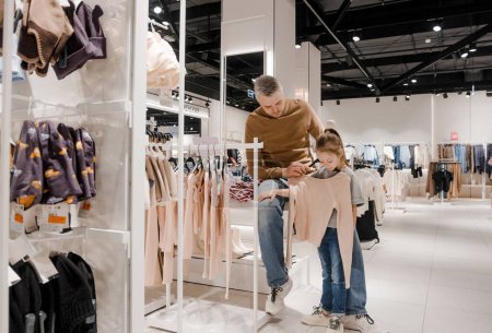 Téléchargez les photos : Un père aide sa jeune fille à choisir des vêtements dans un magasin de détail moderne et lumineux. - en image libre de droit