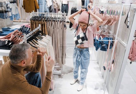 Un père regarde sa fille essayer un chapeau rose et un sac à main à carreaux dans un magasin de vêtements, entouré de supports de vêtements à la mode.