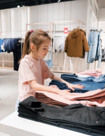 A young girl wearing a pink t-shirt organizing and browsing jeans at a clothing store, surrounded by various apparel items.