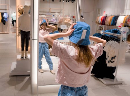 A young woman adjusts a blue hat while looking in a mirror in a brightly lit, modern clothing store.